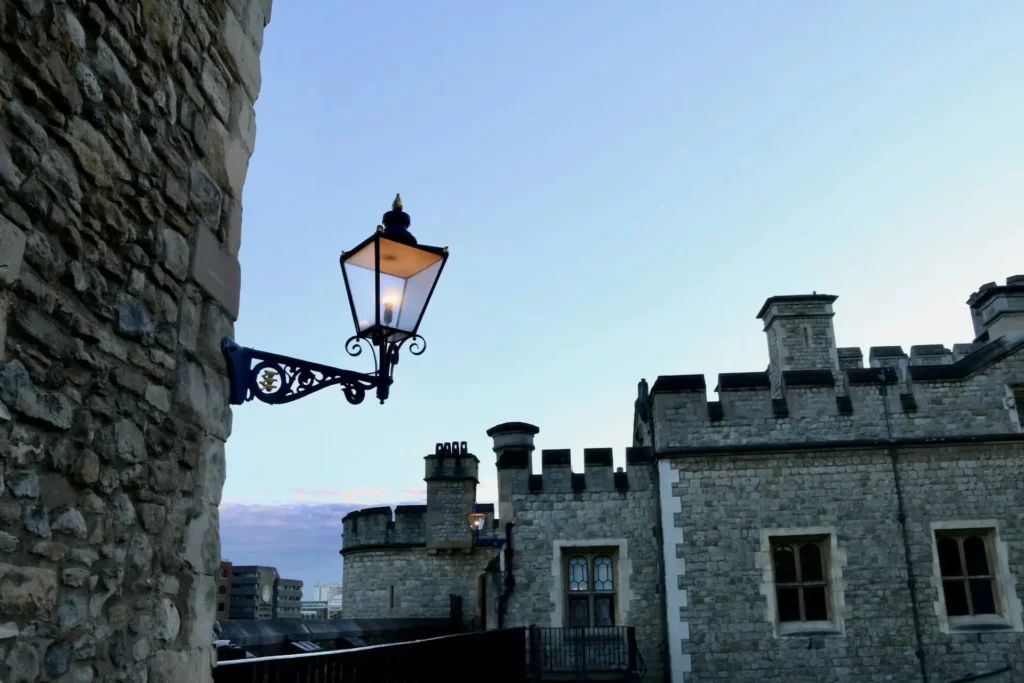 A historic-style lamp attached to a stone wall with a softly glowing light, with a medieval castle in the background at dusk.
