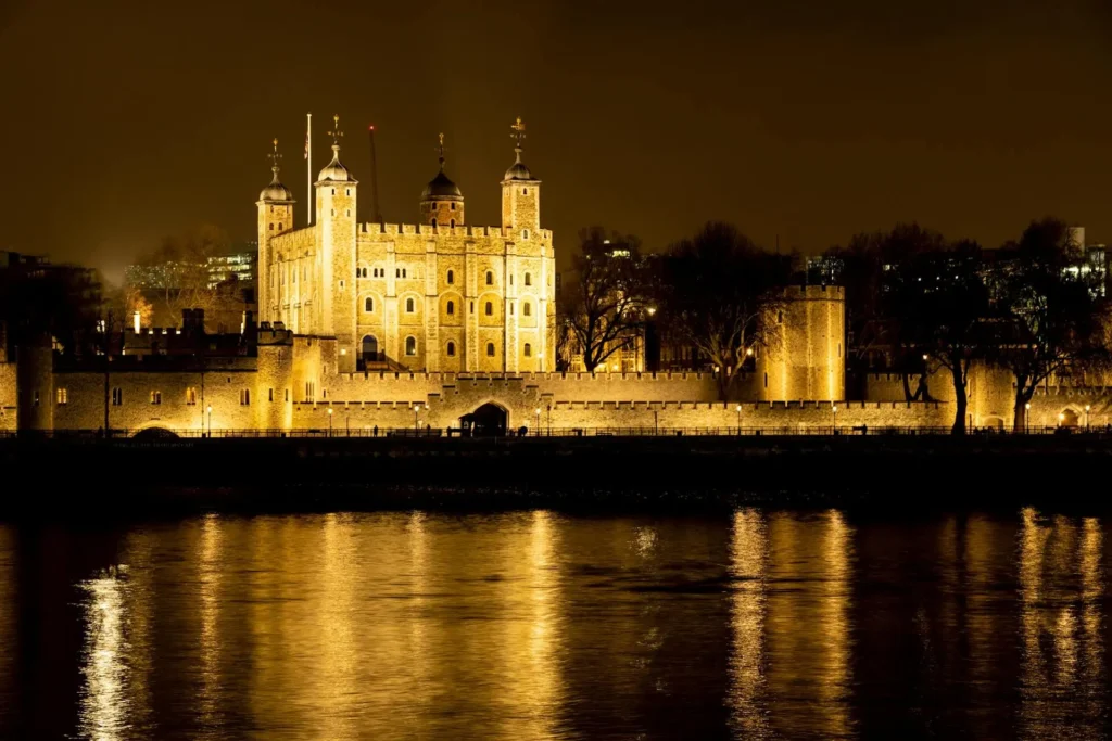 A nighttime view of the Tower of London with golden lights reflecting on the River Thames