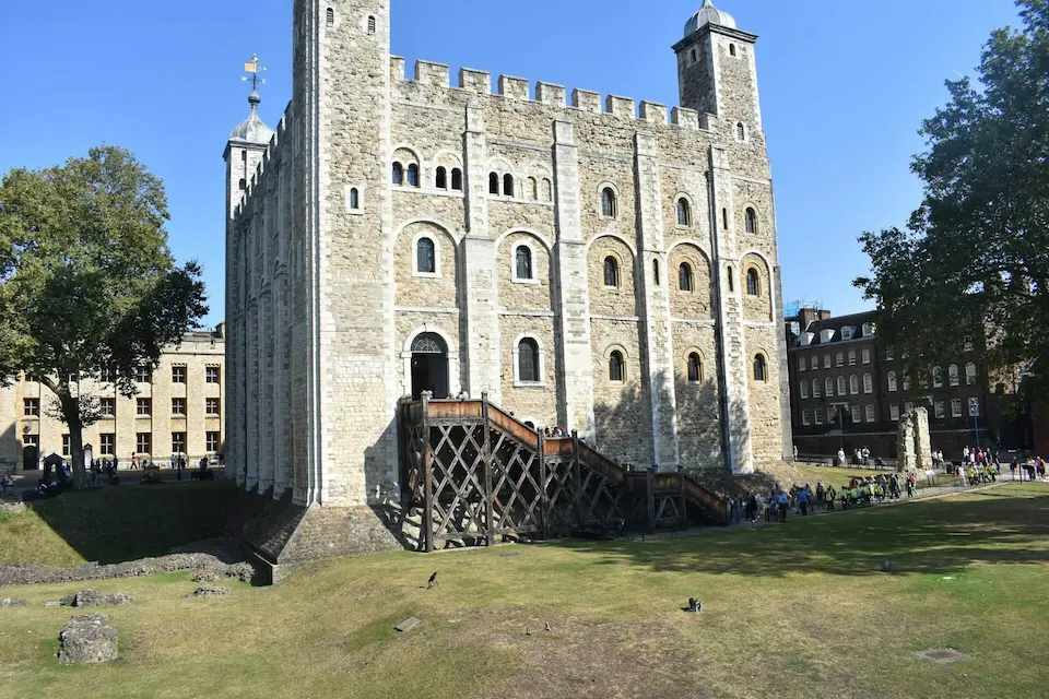 The White Tower, a large Norman stone keep at the Tower of London, standing tall against a blue sky with visitors exploring the area.